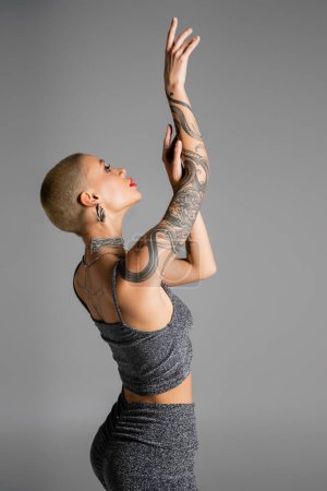 Photo for Side view of trendy woman in lurex crop top posing with raised tattooed hands isolated on grey - Royalty Free Image