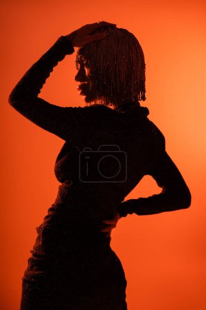 Foto de Silueta sombreada de mujer glamour sexy en vestido negro y ropa de cabeza metálica sobre fondo naranja - Imagen libre de derechos