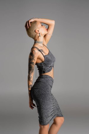 Photo for Side view of sexy tattooed woman in stylish lurex clothes posing with hand above head isolated on grey - Royalty Free Image