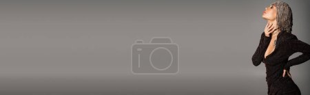 Téléchargez les photos : Vue latérale de femme sensuelle en robe noire et coiffure bijoux brillants touchant le cou isolé sur gris, bannière - en image libre de droit