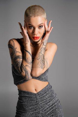 Photo for Seductive tattooed woman with short hair and bright red lips touching head and looking at camera isolated on grey - Royalty Free Image