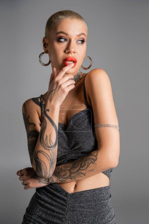 seductive tattooed woman in hoop earrings and lurex crop top touching red lips and looking away isolated on grey