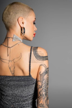 back view of short haired tattooed woman in metal necklaces and shiny top standing isolated on grey