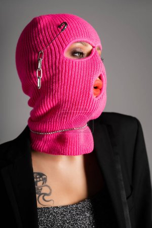 Porträt einer extravaganten Frau in schwarzem Blazer und pinkfarbener Sturmhaube, die isoliert auf grau blickt