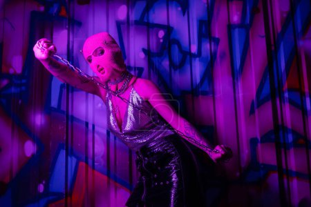 seductora mujer en top metálico y pasamontañas sosteniendo cadena de plata cerca de la pared con coloridos graffiti en luz púrpura