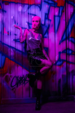 Téléchargez les photos : Pleine longueur de femme élégante en cagoule tenant chaîne d'argent et regardant loin près du mur avec des graffitis dans la lumière violette - en image libre de droit