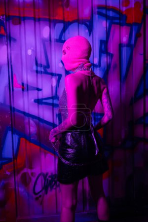 vista trasera de mujer sexy en pasamontañas y falda de cuero negro de pie con cadena de plata cerca de graffiti colorido en luz de neón púrpura
