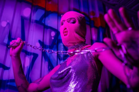 low angle view of sexy woman in pink balaclava and shiny top holding silver chain and looking at camera in purple neon light tote bag #645513934
