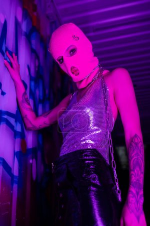 low angle view of provocative tattooed woman in shiny top and pink balaclava posing near graffiti in purple neon light