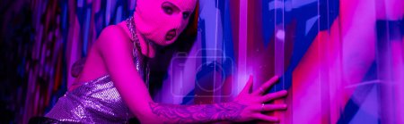 Téléchargez les photos : Femme sexy en cagoule rose regardant la caméra tout en touchant des graffitis colorés en lumière violette, bannière - en image libre de droit