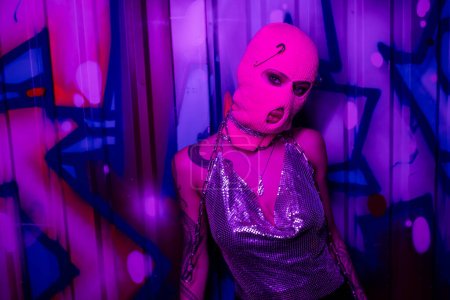 verführerische tätowierte Frau in metallisch glänzendem Oberteil und pinker Sturmhaube, die in Wandnähe steht, mit Graffiti in lila Neonlicht