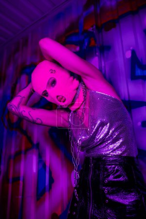 Tiefansicht einer provokanten Frau mit Sturmhaube und metallischem Oberteil, die in lila Licht in die Kamera schaut, in der Nähe bunter Graffiti