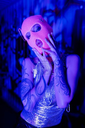 seductive tattooed woman in balaclava and shiny top holding hands near face and looking at camera in blue and purple light