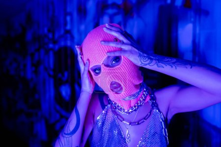 apasionada mujer tatuada en cadenas de cuello plateado mirando a la cámara mientras toca pasamontañas de punto en luz azul y púrpura
