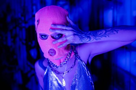 provocativa mujer en pasamontañas y parte superior metálica con collares posando con la mano cerca de la cara en iluminación azul y púrpura