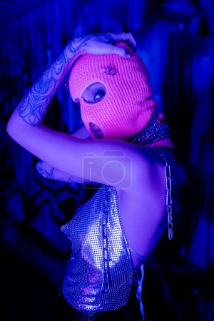 provocativa mujer tatuada en parte superior metálica y pasamontañas de punto mirando a la cámara en luz de neón azul y púrpura