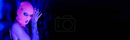anonymous tattooed woman in balaclava looking at camera in blue and purple light near black copy space, banner