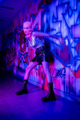 full length of stylish and sexy woman in balaclava looking at camera near wall with graffiti in blue and purple light t-shirt #645514222