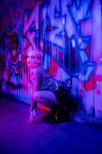 full length of woman in stylish clothes and balaclava sitting near wall with graffiti in blue and purple lighting hoodie #645514262