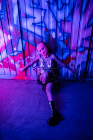 high angle view of passionate woman in balaclava and glamour outfit sitting near colorful graffiti in blue and purple lighting 