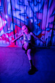 high angle view of passionate woman in balaclava and glamour outfit sitting near colorful graffiti in blue and purple lighting  Tank Top #645514294