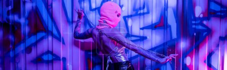 side view of glamour woman in silver top and balaclava posing with chain near colorful graffiti in blue neon light, banner Stickers 645514332