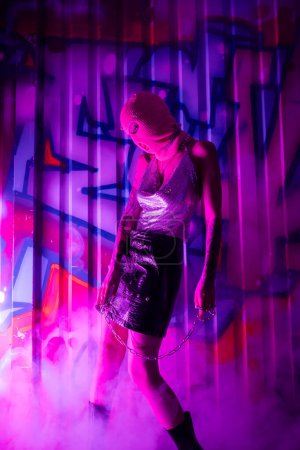 sexy Frau in Sturmhaube und glänzendem Top mit Lederrock, stehend mit Kette neben buntem Graffiti in lila Rauch