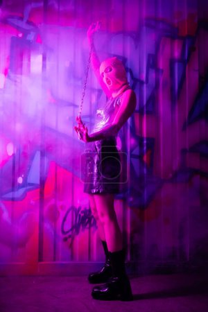 Frau in Sturmhaube und Lederstiefeln posiert mit Kette in der Nähe bunter Graffiti in lila Licht mit Rauch