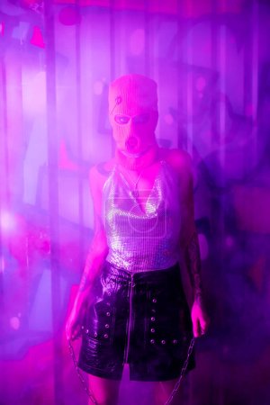 sexy woman in balaclava and silver top holding chain and looking at camera near wall with graffiti in purple light with smoke