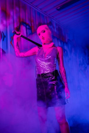 sexy woman in pink balaclava and silver top with leather skirt holding baseball bat near wall with graffiti in purple fog