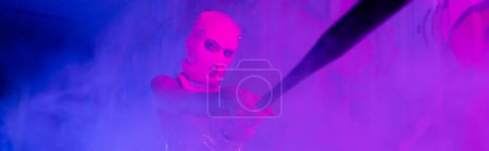 anonymous woman in balaclava holding baseball bat and looking at camera in foggy purple light, banner