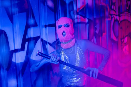 provocative woman in balaclava and silver top grimacing with baseball bat near graffiti in blue and purple light