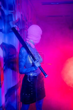 sexy woman in balaclava standing with baseball bat in purple light and looking at pink smoke