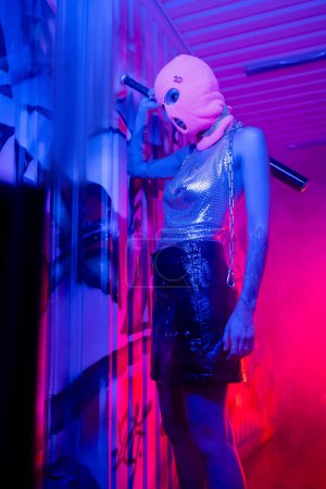 sexy woman in silver top and balaclava standing with baseball bat near wall with graffiti in blue neon light with pink smoke