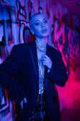 fashionable short haired woman in black blazer and silver necklaces looking at camera near graffiti in blue light with pink smoke tote bag #645514498