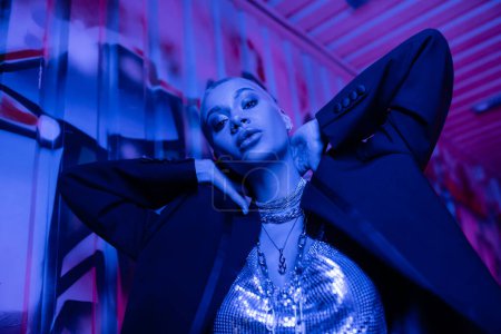 low angle view of extravagant woman in silver top and black jacket posing with hands behind neck near graffiti in blue neon lighting