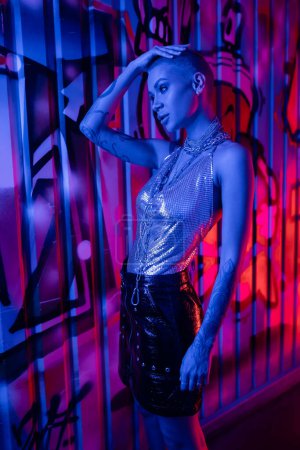 stylish woman in silver top and black leather skirt touching shirt hair near colorful graffiti in blue neon light