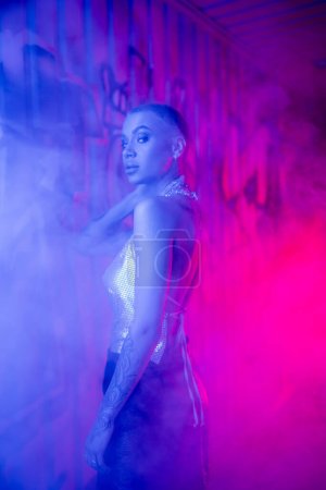 seductive short haired woman in silver top looking at camera near wall with graffiti in blue and pink light with smoke
