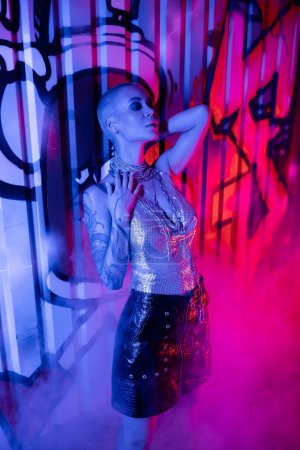 Téléchargez les photos : Femme tatouée passionnée en haut métallique et jupe en cuir noir debout près de graffitis colorés dans la lumière bleue et rose avec de la fumée - en image libre de droit