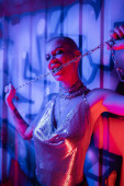 passionate woman in shiny top biting silver chain near wall with graffiti in blue and pink lighting puzzle #645514654