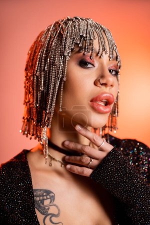 Photo for Portrait of sexy tattooed woman in metallic wig with rhinestones touching neck and looking at camera on coral pink background - Royalty Free Image