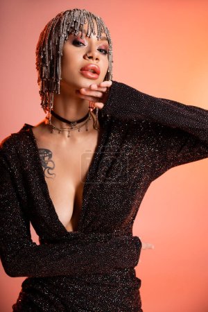 Téléchargez les photos : Femme tatouée avec corps sexy toucher menton tout en posant dans des vêtements glamour sur fond rose corail - en image libre de droit