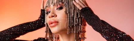 portrait of sexy young woman touching metallic wig with rhinestones and looking at camera on coral pink background, banner