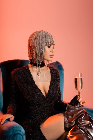 extravagante Frau in schwarzem Kleid und metallischer Kopfbedeckung sitzt im Sessel mit einem Glas Champagner auf korallenrosa Hintergrund
