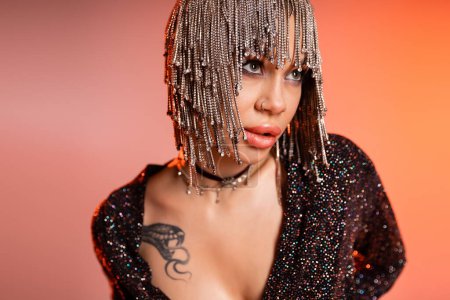 portrait of passionate tattooed woman in metallic headwear with shiny rhinestones looking away on pink and orange background