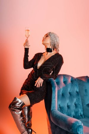 Foto de Mujer en peluca plateada y vestido de lurex negro de pie con copa de champán cerca de sillón de terciopelo sobre fondo rosa y naranja - Imagen libre de derechos
