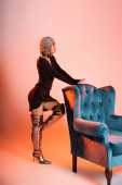 side view of extravagant woman in black dress and long boots posing near velour armchair on pink and orange background Longsleeve T-shirt #645514840