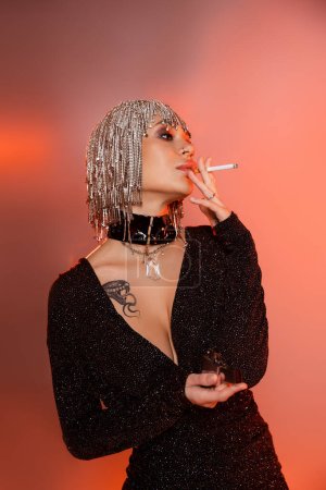 Photo for Tattooed woman in shiny wig and black sexy dress smoking cigarette on pink and orange background - Royalty Free Image