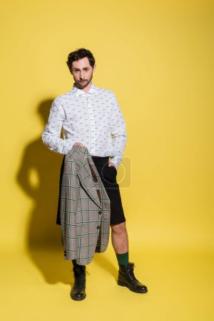 Foto de Longitud completa del hombre de moda en camisa y pantalones cortos que sostienen la chaqueta sobre fondo amarillo - Imagen libre de derechos