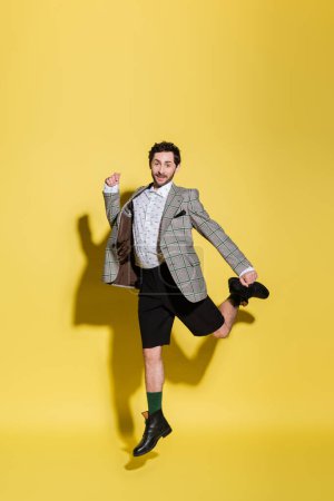 Modelo moderno y sonriente en pantalones cortos y chaqueta saltando sobre fondo amarillo 